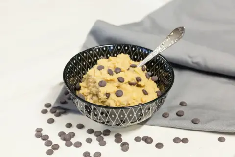 Schüssel mit essbarem Keksteig und Schokostückchen, daneben verstreute Schokotropfen. Ein Löffel steckt im Teig.