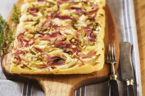 Flammkuchen mit roten Zwiebeln, Pinienkernen und Käse auf einem Holzbrett, daneben Gabel und Messer.