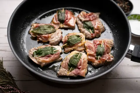 Saltimbocca alla romana in einer Pfanne, umgeben von Kräutern und Gewürzen.