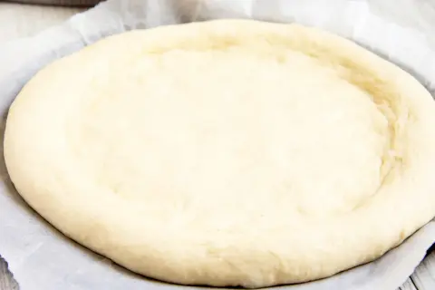 Ein runder, heller pizzateig mit trockenhefe liegt auf Backpapier. Die Ränder sind leicht angehoben.
