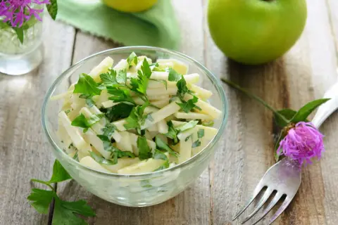 Frischer Kohlrabi Salat mit Petersilie in einer Glasschüssel, garniert mit einer lila Blüte und grünen Äpfeln im Hintergrund.