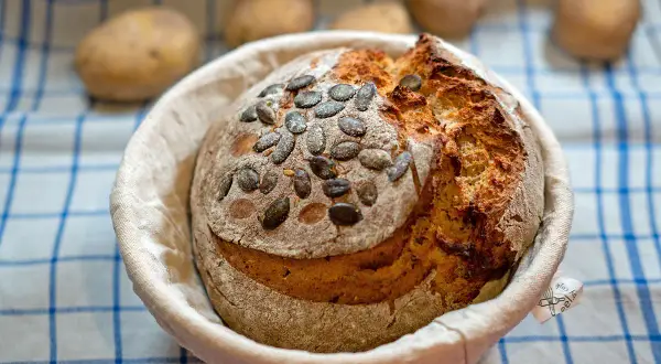 Ein frisch gebackenes Kartoffelbrot mit Kürbiskernen, bereit für das Kartoffelbrot Rezept. Kartoffeln liegen unscharf im Hintergrund.