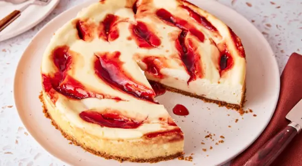 Ein cremiger american cheesecake mit roten Fruchtsaucen-Wirbeln auf einem Teller, bereit zum Genießen.