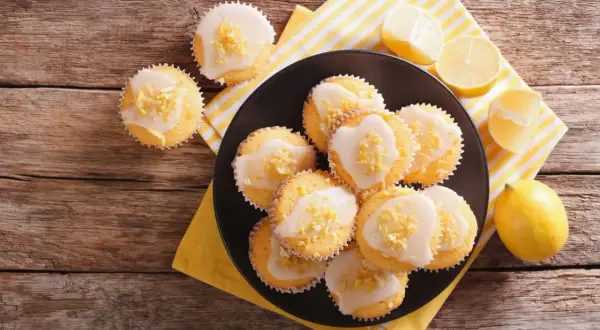 Zitronen-Muffins, frisch aus dem Thermomix, mit Zuckerguss und Zitronenschale bestreut, auf einem Teller mit Zitronenspalten.