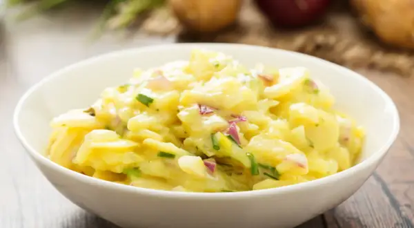 Ein Schüsselchen mit Omas Kartoffelsalat, cremig und mit frischen Kräutern verfeinert. Dazu liegen Kartoffeln und Zwiebeln bereit.