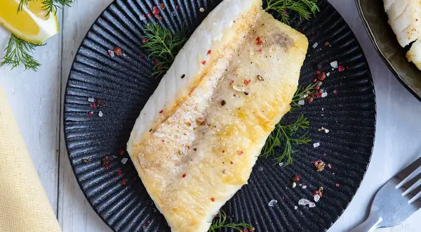 Perfekt gebratenes Rotbarschfilet auf schwarzem Teller, garniert mit Dill und bunten Pfefferkörnern. Dazu Zitronenspalten.