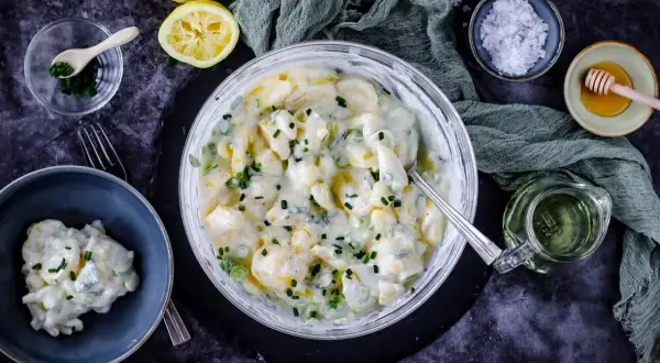 Cremiger Kartoffelsalat mit Joghurt, garniert mit Schnittlauch. Dazu Zitronenspalten und eine kleine Schüssel mit Kräutern.