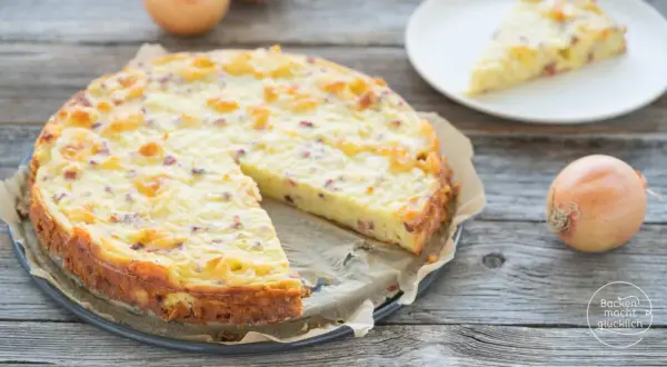 Ein goldbrauner Zwiebelkuchen ohne Boden, mit Speckstücken und einer cremigen Füllung, liegt auf einem Holztisch. Daneben eine Scheibe auf einem Teller und eine Zwiebel.