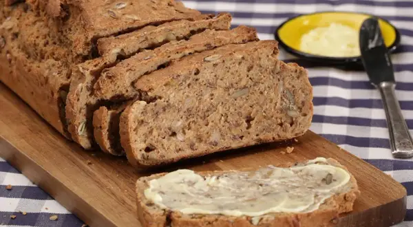 Ein frisch gebackenes Vollkornbrot mit vielen Kernen, aufgeschnitten auf einem Holzbrett. Ein Stück ist mit Butter bestrichen. Perfekt für ein gesundes Frühstück oder Abendbrot.