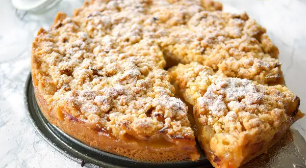 Ein goldbrauner Apfelkuchen mit Streuseln, bestäubt mit Puderzucker. Ein Stück wird gerade herausgehoben.