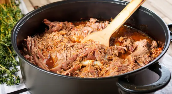 Zartes pulled pork im Dutch Oven, bereit zum Servieren. Ein Holzlöffel liegt darin.