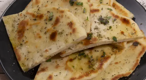 Griechisches Fladenbrot mit Trockenhefe, bestreut mit Kräutern und Olivenöl, auf einem schwarzen Teller.