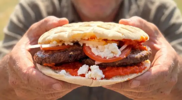 Ein Mann hält eine saftige Pljeskavica mit Tomaten, Zwiebeln und Käse. Ein tolles Rezept für den Sommer!