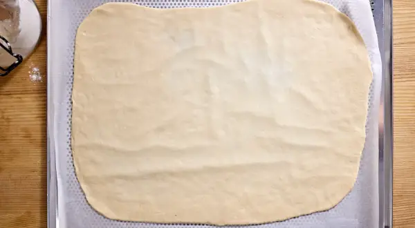 Dünn ausgerollter Flammkuchenteig liegt auf einem Backblech, bereit für leckere Beläge.