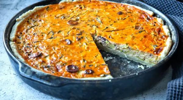Eine köstliche Zucchini-Quiche mit Oliven in einer Gusseisenpfanne, eine Scheibe herausgeschnitten.