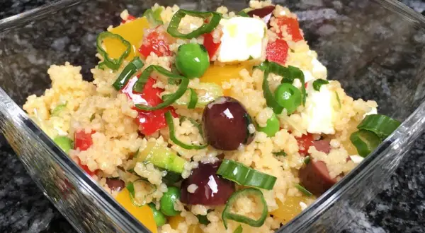 Frischer Couscous Salat mediterran mit Oliven, Paprika, Erbsen und Feta in einer Glasschüssel.