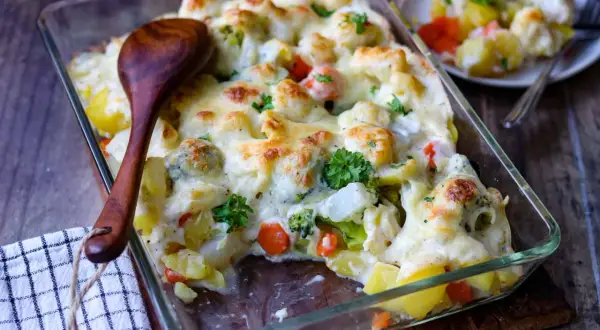 Herzhafter vegetarischer Auflauf mit Kartoffeln, Brokkoli, Blumenkohl und Karotten, überbacken mit Käse. Ein Holzlöffel liegt bereit.