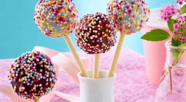 Bunte cake pops mit Schokoglasur und Streuseln in einer weißen Vase, bereit für eine fröhliche Feier.