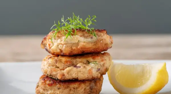 Gestapelte Fischbuletten, garniert mit Dill, neben einer Zitronenspalte. Ein leckeres Buletten Rezept.