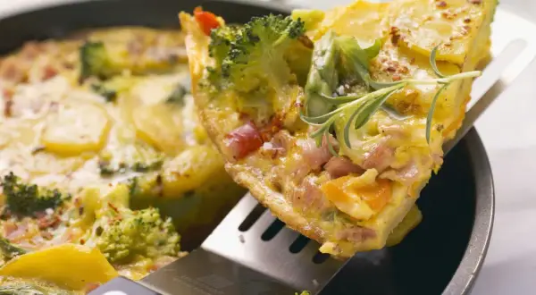 Ein Stück herzhaftes Omelett mit Brokkoli, Kartoffeln und Schinken wird aus einer Pfanne gehoben. Ein tolles Omelett Rezept für jeden Tag.