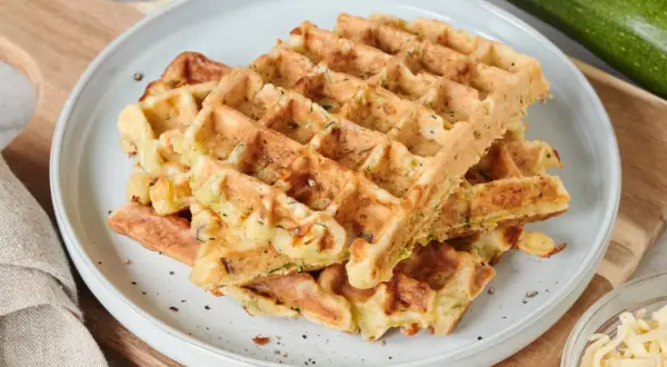Goldbraune Zucchini-Waffeln auf einem Teller, daneben frische Zucchini und geriebener Käse. Ein leckeres zucchini waffeln rezept.