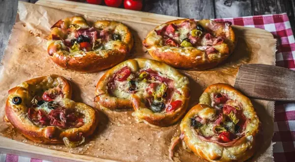 Fünf kleine Brezel Pizza mit Schinken, Oliven und Tomaten, auf Backpapier auf einem Holzbrett angerichtet.