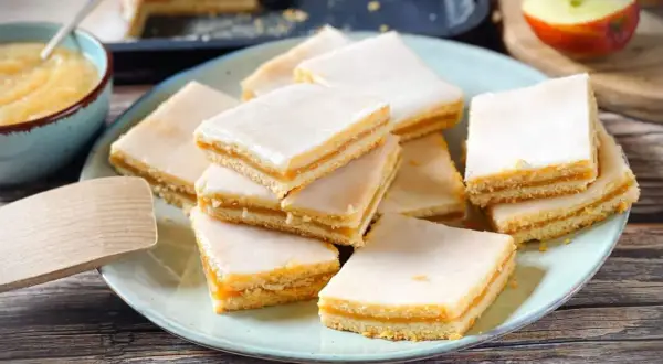 Rechteckige Stückchen Apfelkuchen mit einer zarten Apfelmusfüllung und weißer Glasur, auf einem Teller angerichtet.