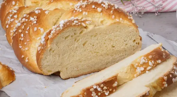 Ein goldbrauner Hefezopf, bestreut mit Zuckerkristallen, liegt auf Backpapier. Ein Stück ist angeschnitten, daneben liegen drei Scheiben. Ein tolles Rezept für Hefezopf!