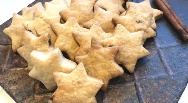 Ein Haufen goldbrauner Dinkelkekse ohne Zucker in Sternform, daneben Zimtstangen auf einem rustikalen Teller.