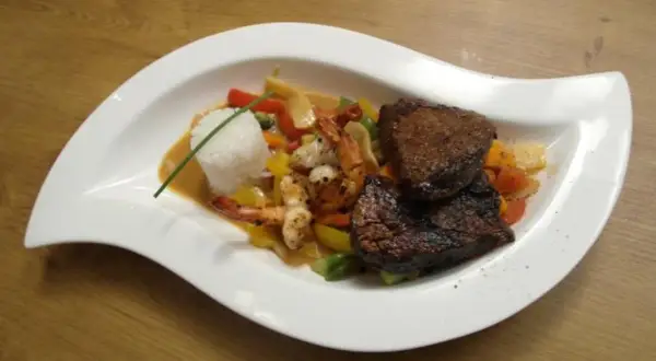 Zwei saftige Rinderfiletstücke auf einem Bett aus Gemüse und Garnelen, serviert mit Reis in einer cremigen Sauce, die an einen **thai curry** erinnert.