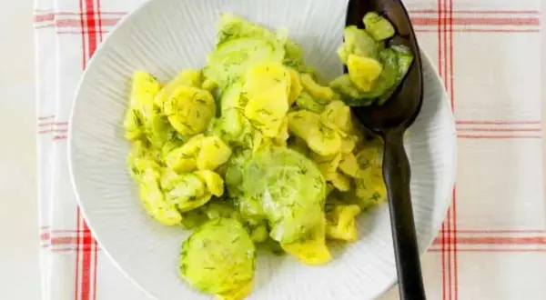 Ein Teller mit Kartoffelsalat mit Gurken und Dill, garniert mit frischem Dill. Ein Löffel mit dem Salat liegt auf dem Teller.