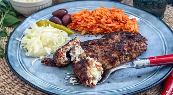 Gefülltes Bifteki-Rezept auf einem Teller mit Reisnudeln, Krautsalat, Oliven und Peperoni. Ein Stück wird mit einer Gabel angeschnitten.