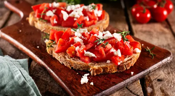 Zwei rustikale Scheiben Brot belegt mit frischen Tomatenwürfeln und zerbröseltem Feta. Bruschetta mit Feta, garniert mit Kräutern, auf einem Holzbrett.