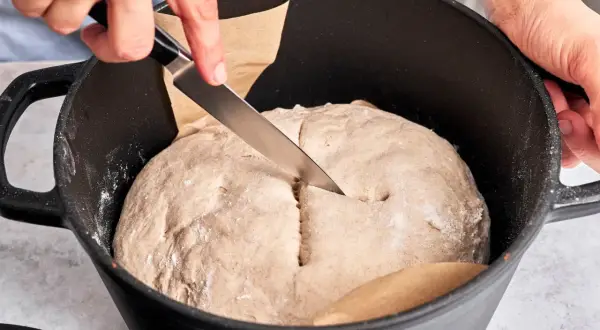 Hände schneiden mit Messer in den Teig für ein "friss dich dumm Brot" in einem Gusseisentopf.