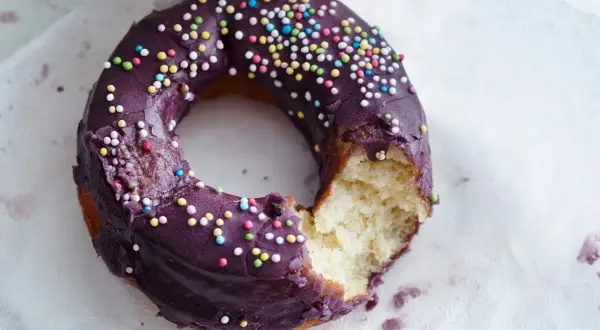 Ein lila glasierter Donut mit bunten Streuseln, ein Bissen fehlt. Perfekt für ein schnelles Donuts Rezept.