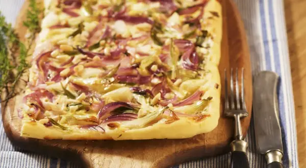 Flammkuchen mit roten Zwiebeln, Pinienkernen und Käse auf einem Holzbrett, daneben Gabel und Messer.