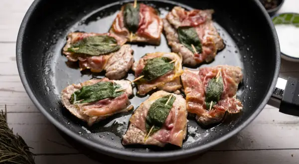 Saltimbocca alla romana in einer Pfanne, umgeben von Kräutern und Gewürzen.