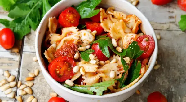 Schüssel mit Nudelsalat, Farfalle, Kirschtomaten, Mozzarella, Pinienkernen und Rucola. Ein köstlicher nudelsalat mit getrockneten tomaten.