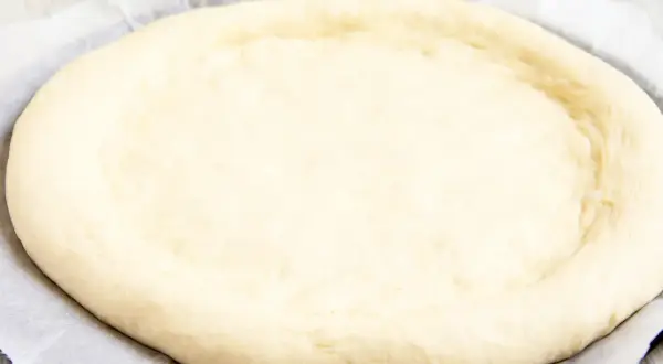 Ein runder, heller pizzateig mit trockenhefe liegt auf Backpapier. Die Ränder sind leicht angehoben.