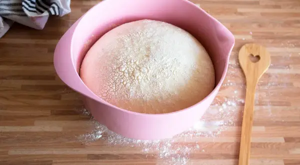 Hefeteig für einen Blechkuchen ruht in einer rosa Schüssel. Daneben liegt ein Holzlöffel mit Herz.