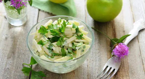Frischer Kohlrabi Salat mit Petersilie in einer Glasschüssel, garniert mit einer lila Blüte und grünen Äpfeln im Hintergrund.