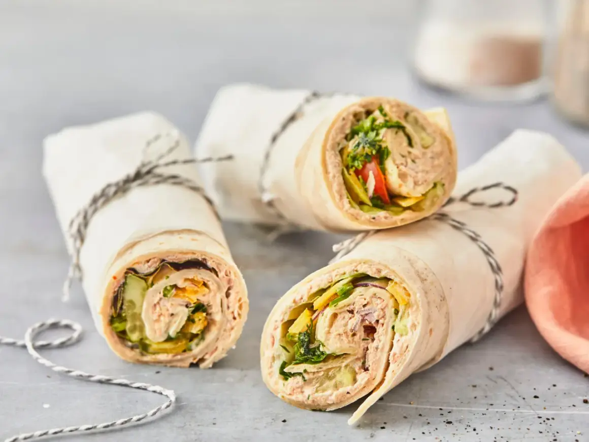Drei leckere Thunfisch-Wraps, gefüllt mit Salat, Gurke und Tomate, sind in Papier gewickelt und mit Schnur verschnürt.