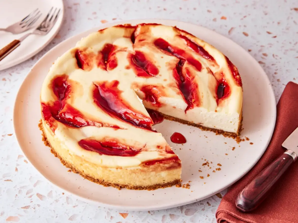 Ein cremiger american cheesecake mit roten Fruchtsaucen-Wirbeln auf einem Teller, bereit zum Genießen.