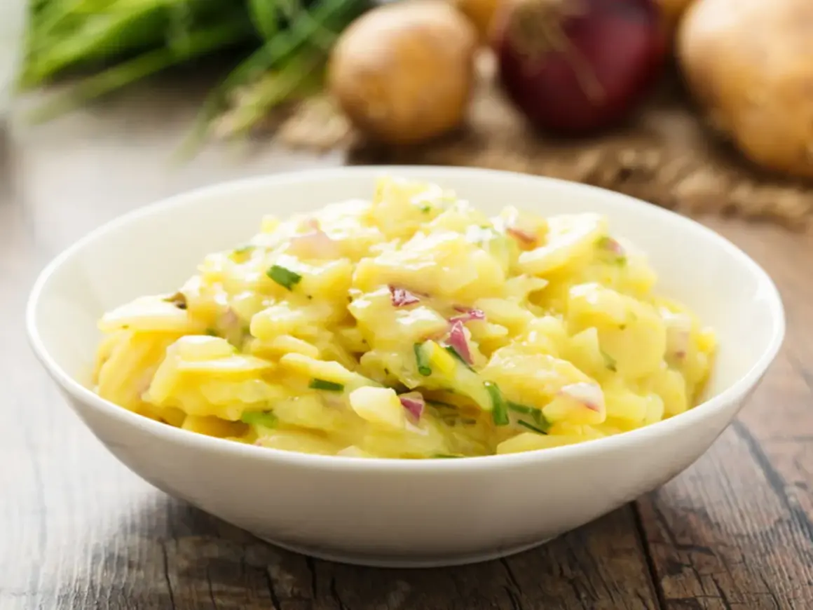 Ein Schüsselchen mit Omas Kartoffelsalat, cremig und mit frischen Kräutern verfeinert. Dazu liegen Kartoffeln und Zwiebeln bereit.