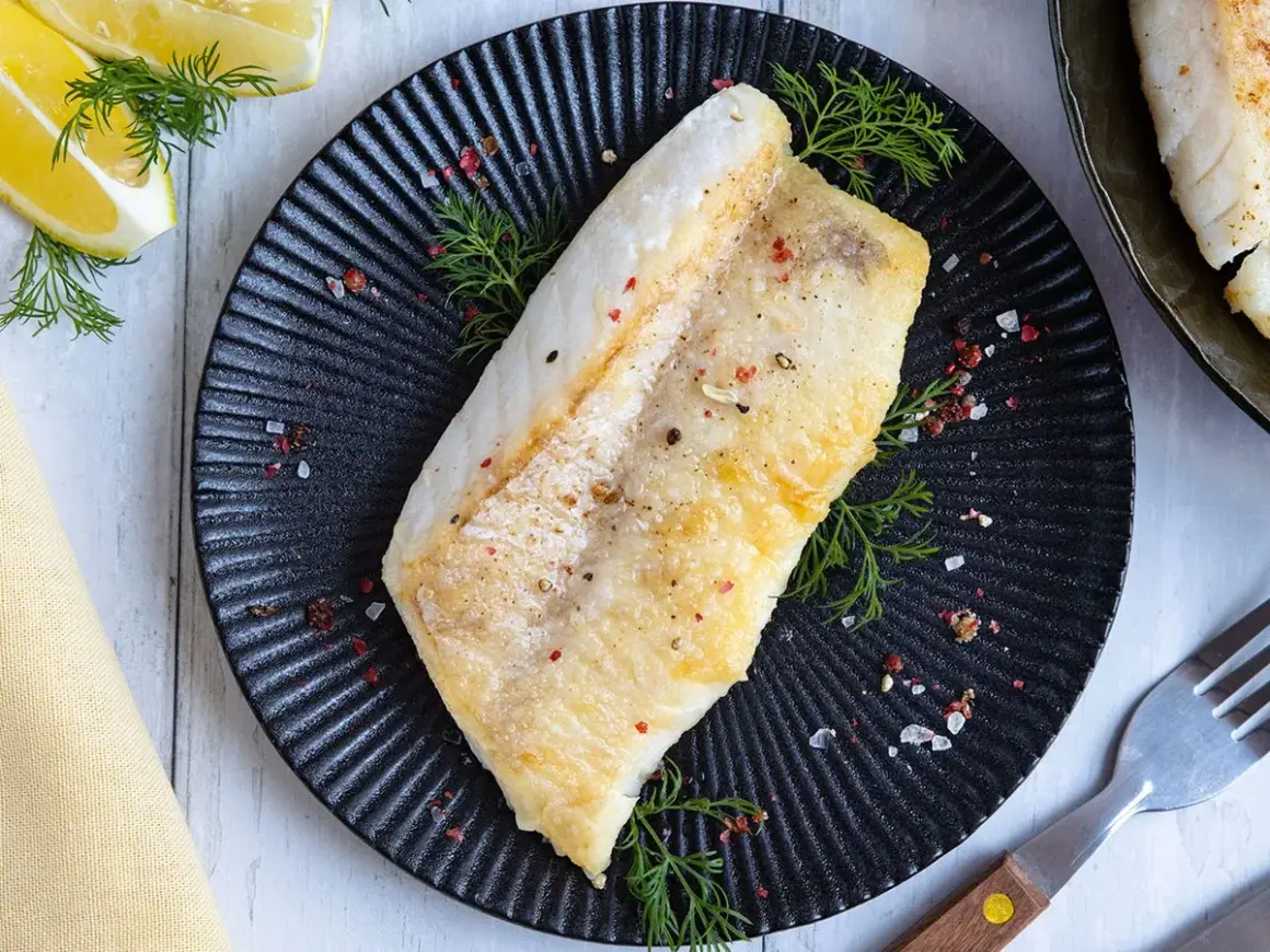 Perfekt gebratenes Rotbarschfilet auf schwarzem Teller, garniert mit Dill und bunten Pfefferkörnern. Dazu Zitronenspalten.