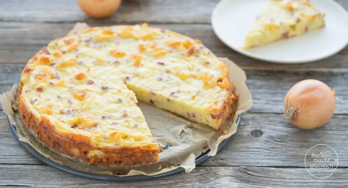 Ein goldbrauner Zwiebelkuchen ohne Boden, mit Speckstücken und einer cremigen Füllung, liegt auf einem Holztisch. Daneben eine Scheibe auf einem Teller und eine Zwiebel.
