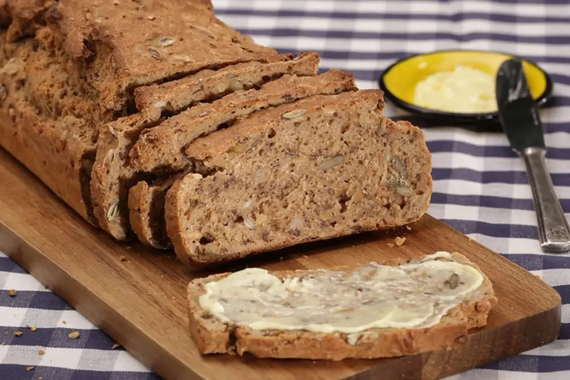 Ein frisch gebackenes Vollkornbrot mit vielen Kernen, aufgeschnitten auf einem Holzbrett. Ein Stück ist mit Butter bestrichen. Perfekt für ein gesundes Frühstück oder Abendbrot.