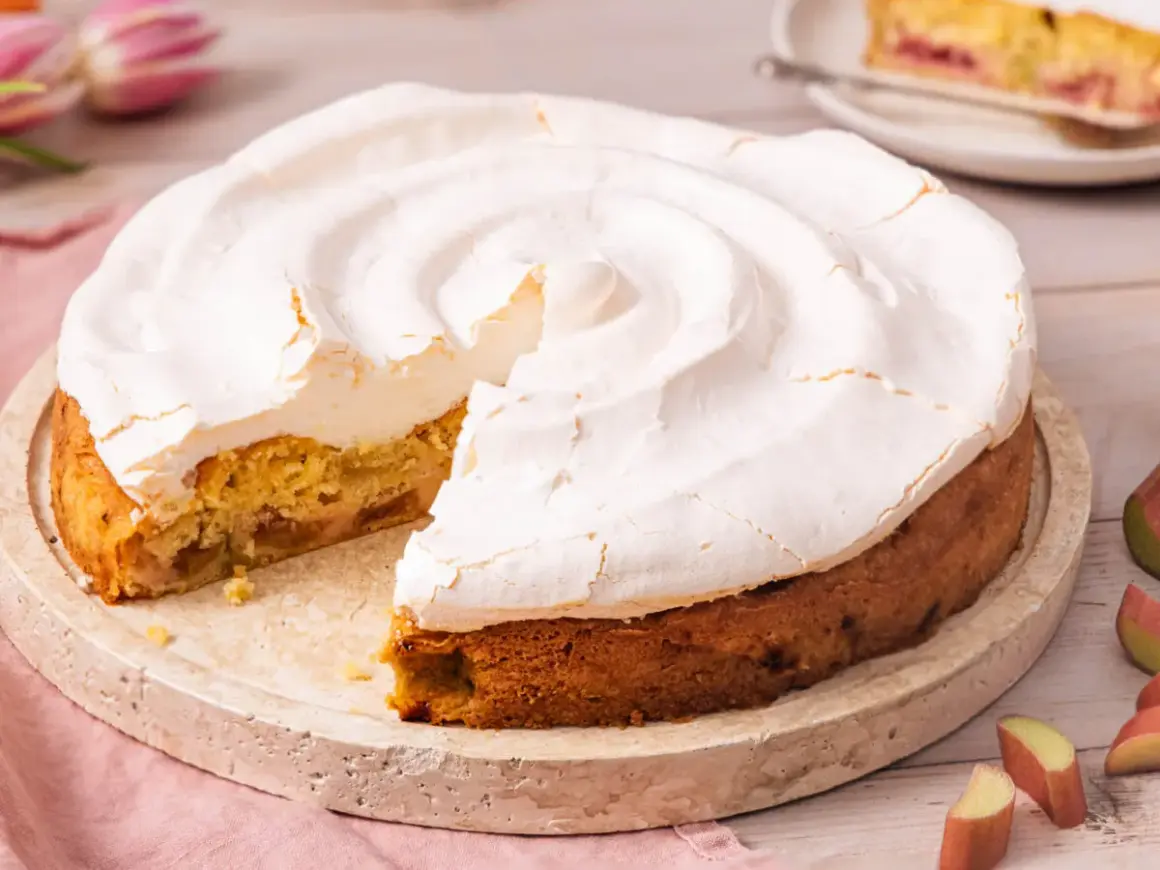 Ein köstlicher Rhabarberkuchen mit Baiser, dessen fluffige Haube leicht gebräunt ist. Ein Stück fehlt bereits.
