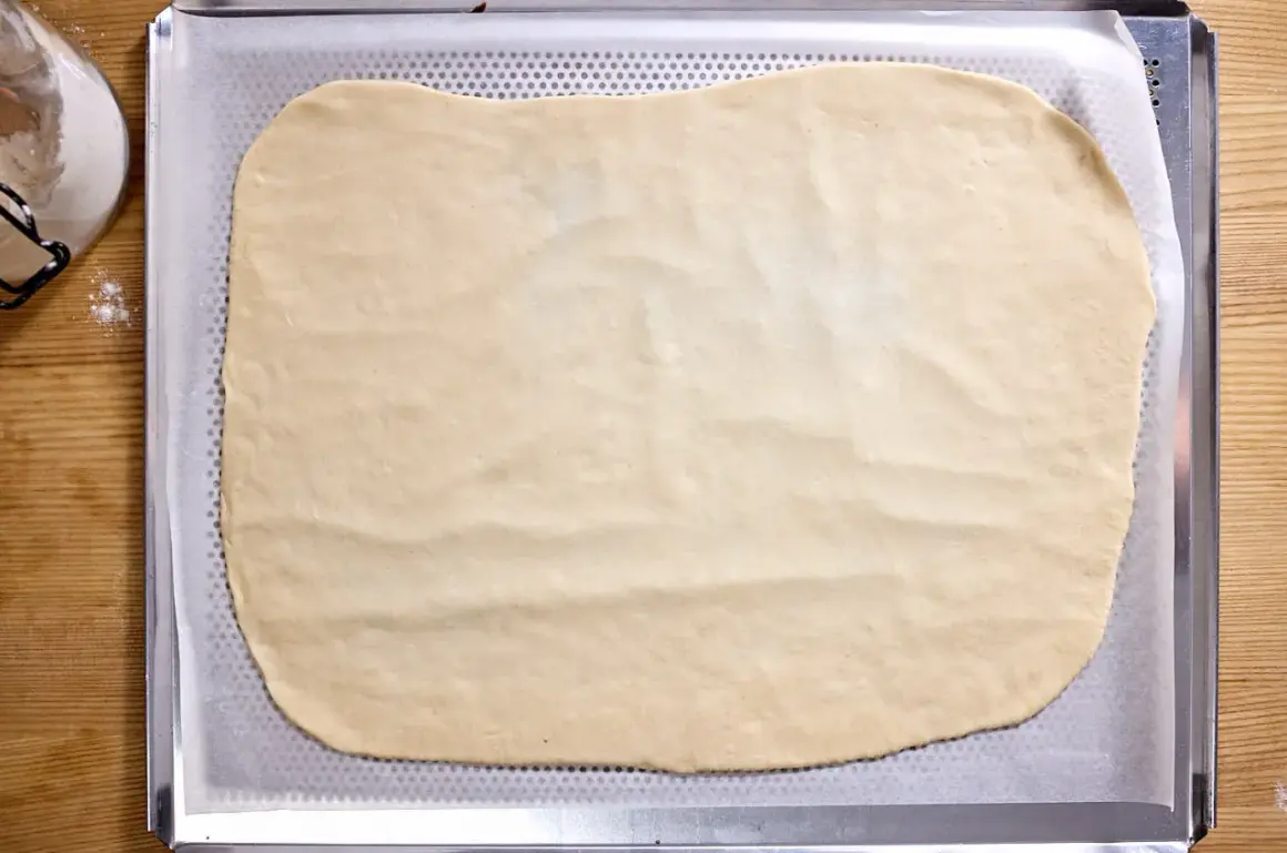 Dünn ausgerollter Flammkuchenteig liegt auf einem Backblech, bereit für leckere Beläge.
