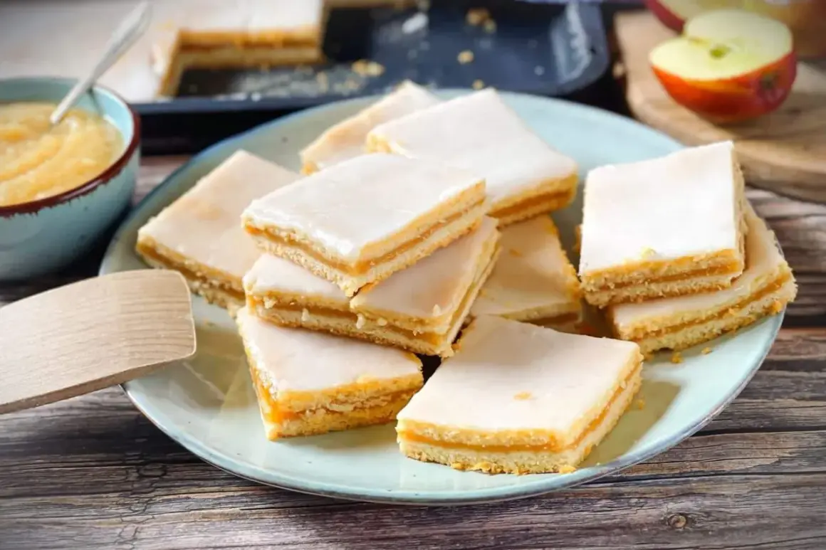 Rechteckige Stückchen Apfelkuchen mit einer zarten Apfelmusfüllung und weißer Glasur, auf einem Teller angerichtet.